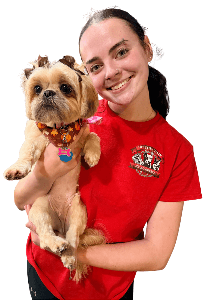 A woman in a red T-shirt holds a small tan dog wearing a decorative collar. Both are facing the camera and smiling.
