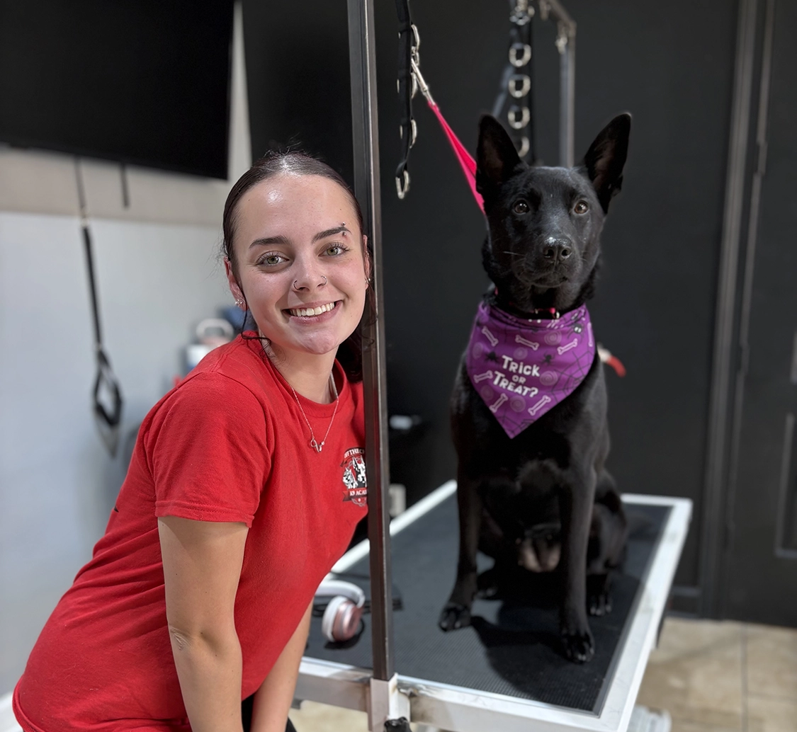 Smiling student in red shirt posing with a black dog wearing a purple bandana on a grooming table, illustrating hands-on training at OTCK9 Grooming Academy.