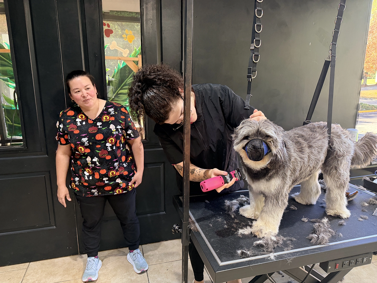 Woman in colorful dog-themed scrubs observing a groomer trimming a dog on a grooming table, with dog wearing a muzzle and fur clippings on the table, highlighting hands-on dog grooming training.