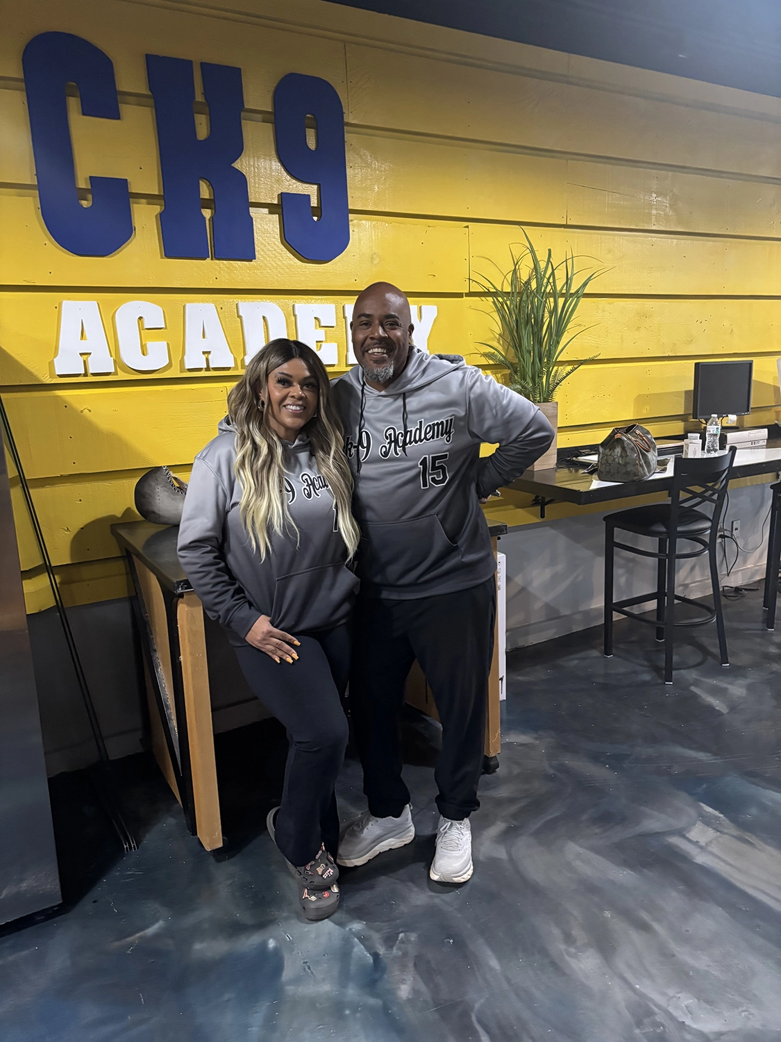 Darrell and Kim, founders of OTCK9 Grooming Academy, standing in front of the academy's logo on a yellow wall, promoting hands-on dog grooming education in Woodstock, GA.