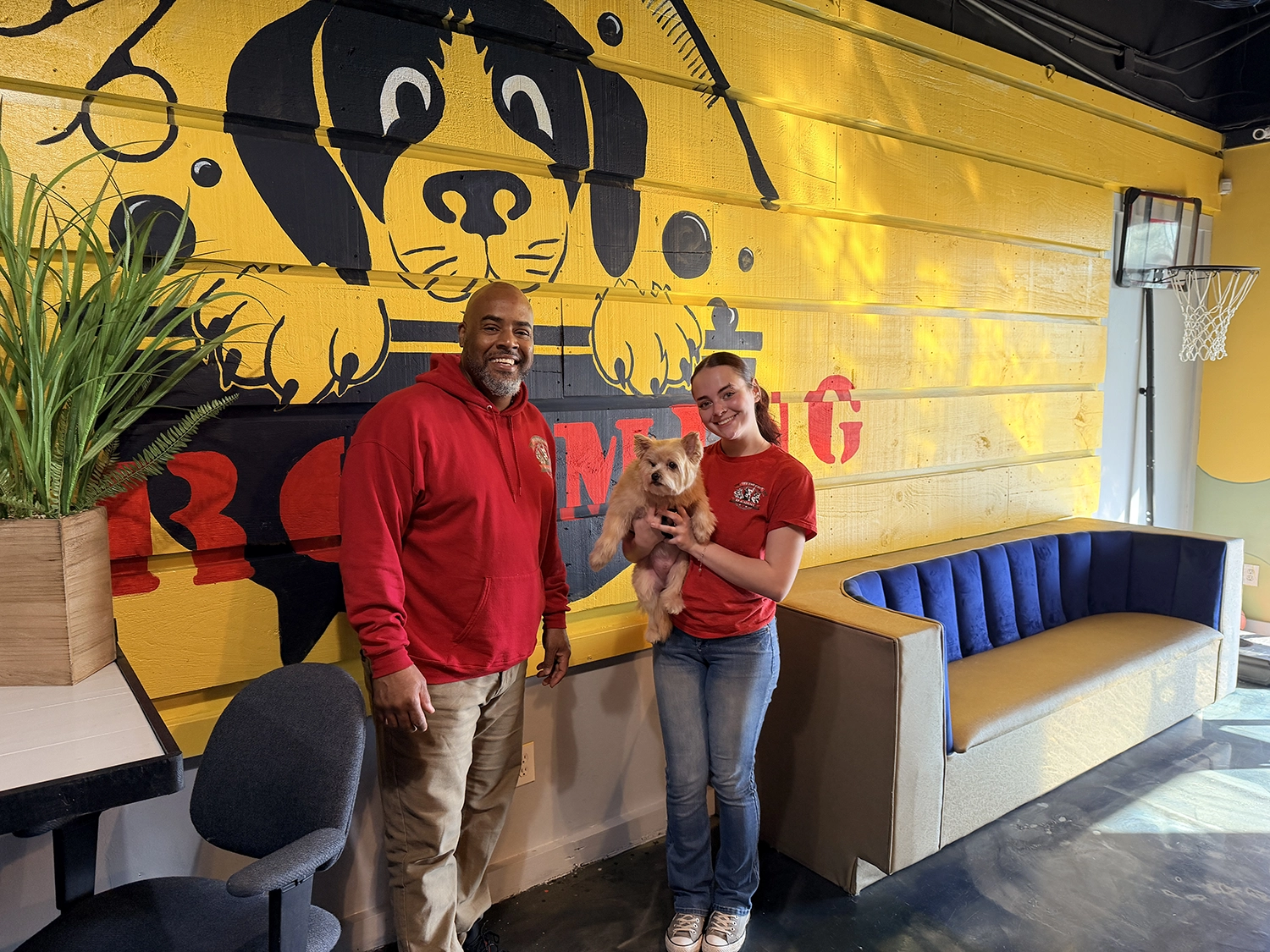Two people stand inside a pet grooming shop, one holding a small dog. A large mural of a dog's face is painted on the yellow wall behind them, adding character to the space and saying much about their love for pets.
