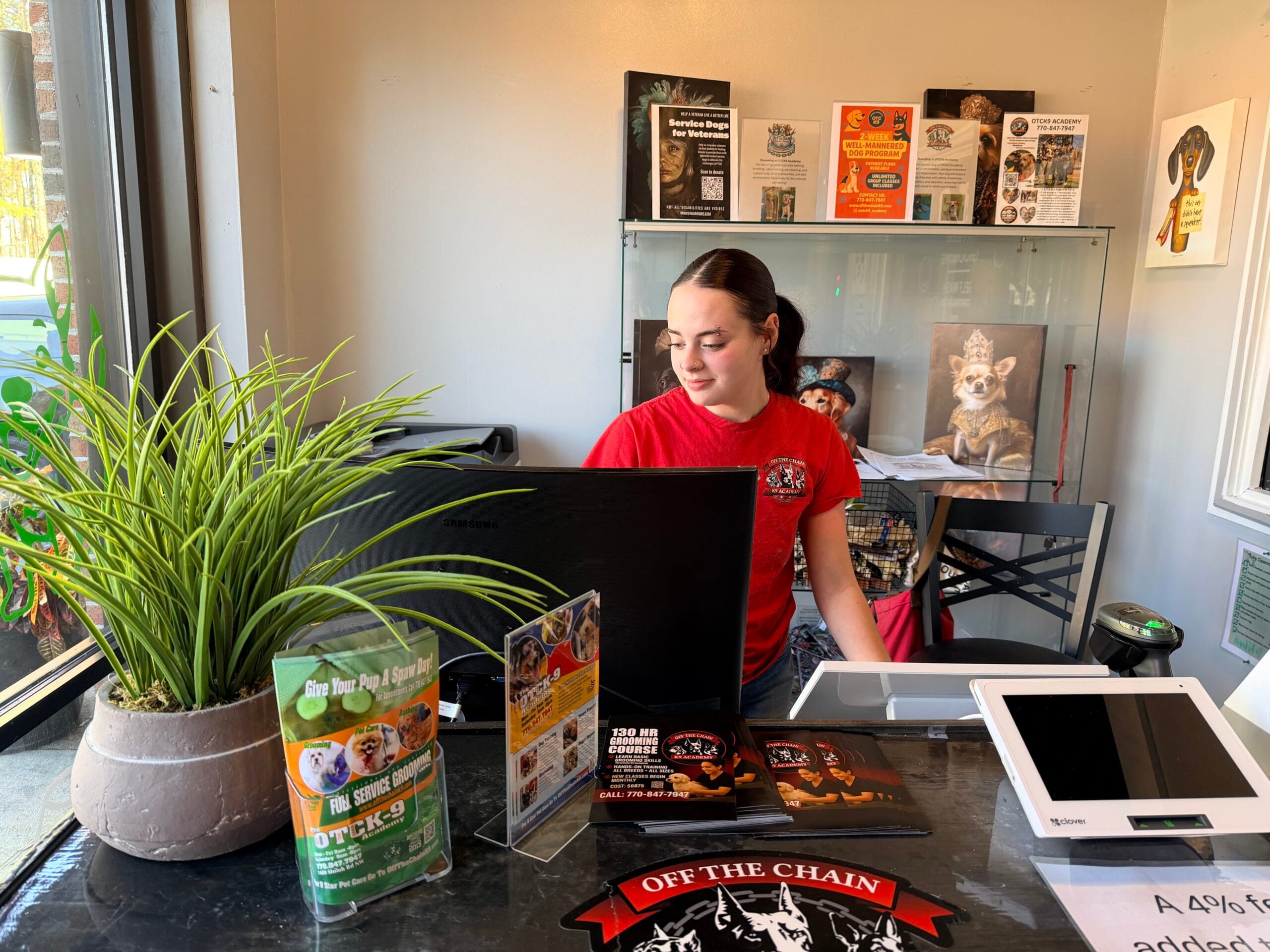 A young woman in a red shirt works at the admissions front desk with a computer, cash register, brochures, and pet-themed decorations.
