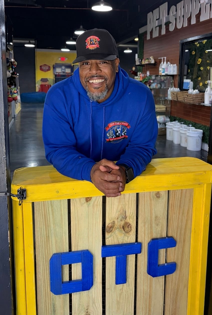 Man in a blue hoodie and black cap smiling and leaning on a wooden counter labeled "OTC" inside a pet supply store, reflecting the positive reviews that make this spot popular with local and global pet lovers.