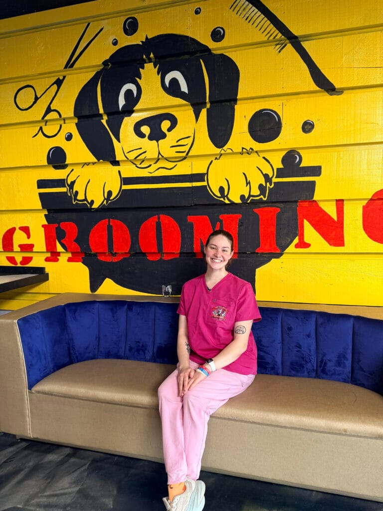 A person in pink scrubs sits on a couch in front of a yellow wall mural of a dog with "GROOMING" written in red letters, highlighting the welcoming atmosphere of the grooming services offered.