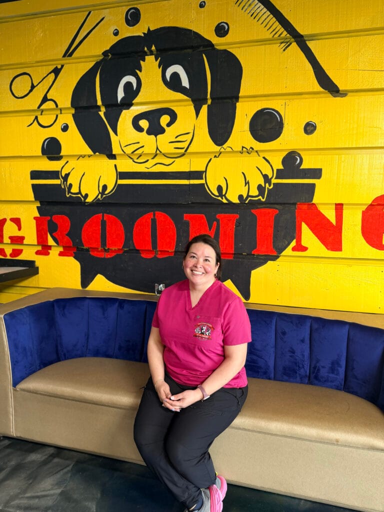 A woman in pink scrubs sits on a blue and tan couch in front of a yellow wall with a large cartoon dog and the words "GROOMING" painted on it, highlighting the welcoming space for grooming services.