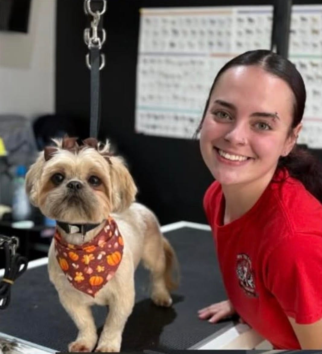A small dog wearing a fall-themed bandana stands on a grooming table next to a smiling woman in a red shirt, showcasing the quality of the grooming services offered.