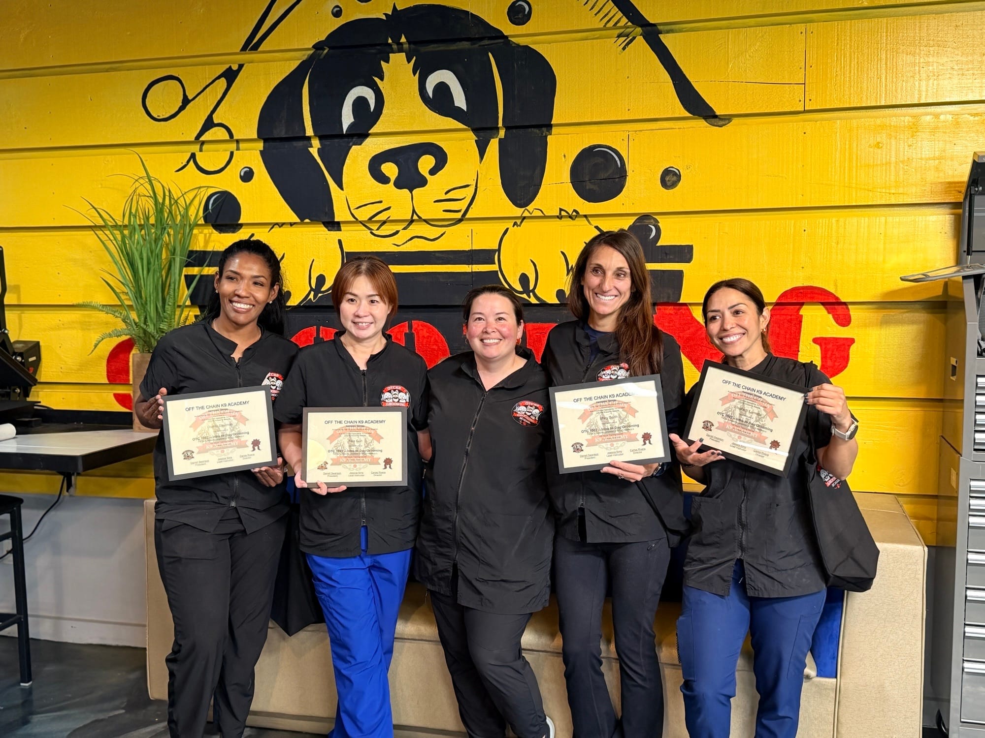 Five women in uniforms stand in front of a yellow wall with a dog graphic, each smiling at the camera while proudly holding their grooming certification.